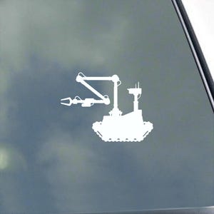 May include: White vinyl sticker depicting a robotic rover with an articulated arm and claw. The rover has a rectangular body with treads and a tall antenna. The sticker is designed for application to a car window or other smooth surface.