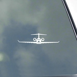 May include: White decal of a private jet, featuring a detailed design of the aircraft's wings, fuselage, and tail. The sticker is applied to a window, with a cloudy sky visible in the background. The jet's design is simple and clean.