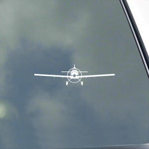 May include: A white vinyl decal featuring a front-facing silhouette of a single-engine propeller airplane. The design displays the aircraft with extended wings, a central propeller, and landing gear, presented as a clean graphic line art illustration.