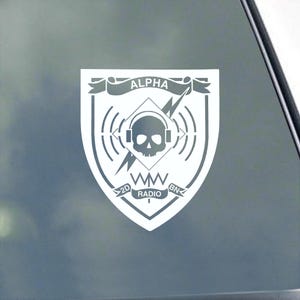May include: White decal featuring a shield design with the word "ALPHA" on a banner. The shield includes a skull wearing headphones, a lightning bolt, and the text "2D RADIO BN".