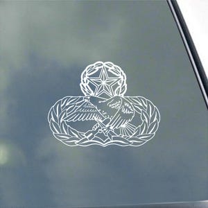 May include: White decal showing a detailed emblem with an eagle holding a staff, encircled by a laurel wreath. Above the eagle is a star-shaped design and a five-pointed star. The decal is likely for a vehicle or window.