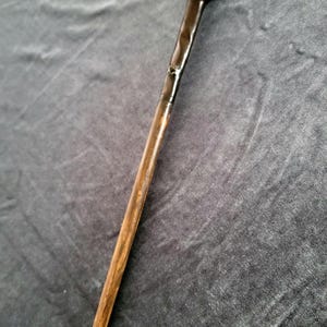 Puede incluir: Una varita mágica de madera hecha a mano con un mango redondeado de color marrón oscuro y un eje cónico de color marrón claro. La varita tiene un acabado brillante y una punta transparente. Es un accesorio o artículo de colección.