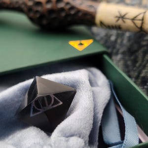 May include: A black, faceted crystal with a white Deathly Hallows symbol, nestled in a light blue velvet pouch inside a green box. A wooden wand is visible in the background.