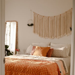 Peut inclure: Une chambre à coucher avec un mur blanc et un lit avec une couette à motifs orange et blanc. Le lit a une tête de lit blanche et un oreiller blanc et orange. Il y a une suspension murale en macramé au-dessus du lit.
