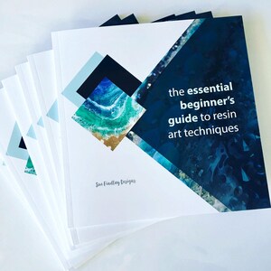May include: A stack of white books with a geometric design and the text "the essential beginner's guide to resin art techniques." The cover art features a blue and green abstract design. The author's name, Sue Findlay Designs, is also printed on the cover.