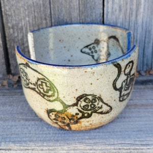 May include: A ceramic bowl with a blue rim and a speckled glaze. The bowl is decorated with a black and white design of video game controllers.