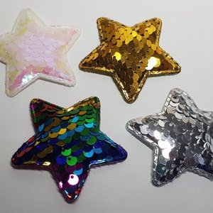 May include: Four star-shaped sequin patches in gold, silver, iridescent white, and a rainbow design. Each star has a stitched border. The patches are set against a white background. Ideal for crafting or embellishing.