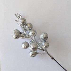 May include: A silver glitter berry stem with multiple round berries. The berries and stem are covered in silver glitter. The stem has a thin black wire at the base. The berries are approximately 2 cm in diameter.