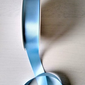 May include: A spool of light blue satin ribbon. The ribbon is wound around a white spool and is partially unwound, showing the soft, silky texture.