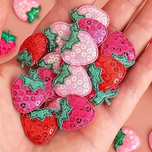 May include: A handful of sequined strawberry patches in pink and red hues, with green glitter leaves. The patches are grouped together, with a few scattered around. These patches are suitable for decorating clothing or accessories.