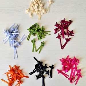 May include: Assortment of satin ribbon rosettes in various colours, including white, green, burgundy, black, orange, and pink. Each rosette is attached to a ribbon tail.