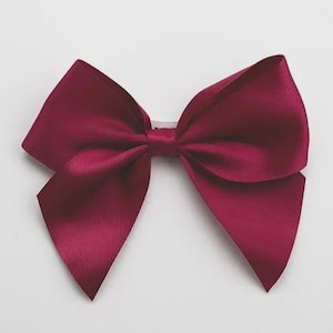 May include: A deep burgundy satin ribbon bow featuring a smooth, lustrous finish and two wide, pointed tails extending from a central knot. The fabric has a subtle sheen that reflects light across its surface, creating a classic, symmetrical shape.