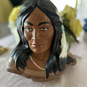 Puede incluir: Un busto de cerámica de una mujer nativa americana con cabello negro largo, piel morena y un collar de cuentas. Lleva un tocado de plumas marrones y blancas.