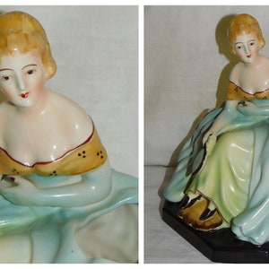 May include: A porcelain figurine of a woman in a blue and green dress, sitting with her arms crossed. The dress has a yellow and brown trim and the woman's hair is blonde. The figurine is a planter with a hole in the back of the dress.
