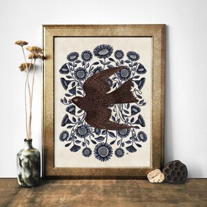 K&ouml;nnte beinhalten: Ein gerahmter Druck eines braunen Vogels, der durch einen Kreis aus blauen Blumen auf cremefarbenem Hintergrund fliegt.