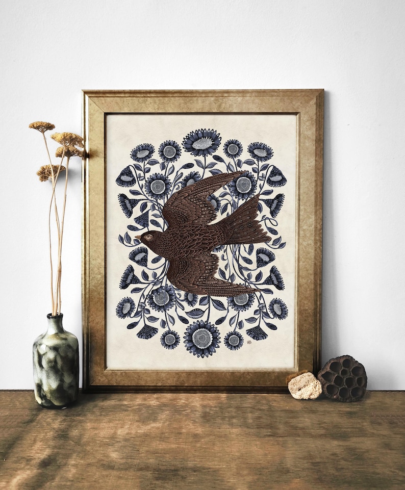 K&ouml;nnte beinhalten: Ein gerahmter Druck eines braunen Vogels, der durch einen Kreis aus blauen Blumen auf cremefarbenem Hintergrund fliegt.