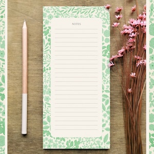 Può includere: Un blocco note bianco con un bordo floreale verde e la parola "NOTES" in alto. Il blocco note è su una superficie di legno con una matita e un mazzo di fiori secchi.