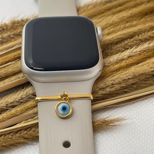 May include: A close-up of a smartwatch with a light gray band and a gold-colored charm. The charm features a blue eye design. The watch face is black.