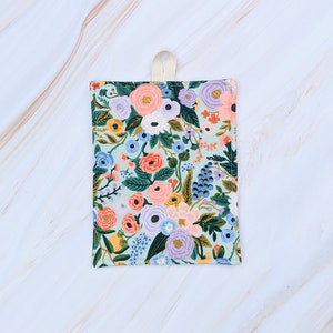 Puede incluir: Un agarrador de olla de tela azul claro con un patrón floral con flores rosas, naranjas, amarillas, moradas y azules. El agarrador de olla tiene un lazo para colgar.