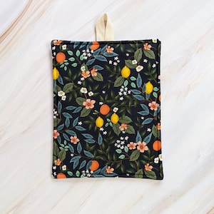 May include: A black cotton pot holder with a floral pattern featuring lemons, oranges, and white flowers. The pot holder has a cream-colored loop for hanging.