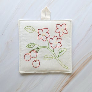 Puede incluir: Un posavasos de algodón blanco con un diseño floral bordado en hilo rojo y verde. El diseño presenta cinco flores y tres cerezas.