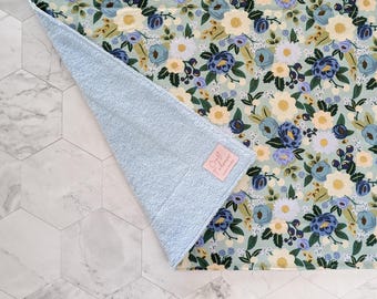 May include: A floral patterned fabric with a light blue terry cloth edge. The fabric features a repeating pattern of blue, white, and yellow flowers on a light green background. A pink label with the text "Craft Tuberose" is visible.