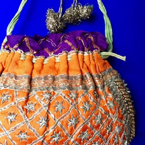 May include: An orange drawstring bag with silver embroidery and a purple lining. The bag has a green and silver cord with tassels.