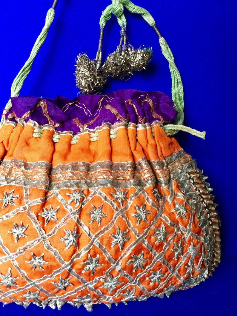 May include: An orange drawstring bag with silver embroidery and a purple lining. The bag has a green and silver cord with tassels.