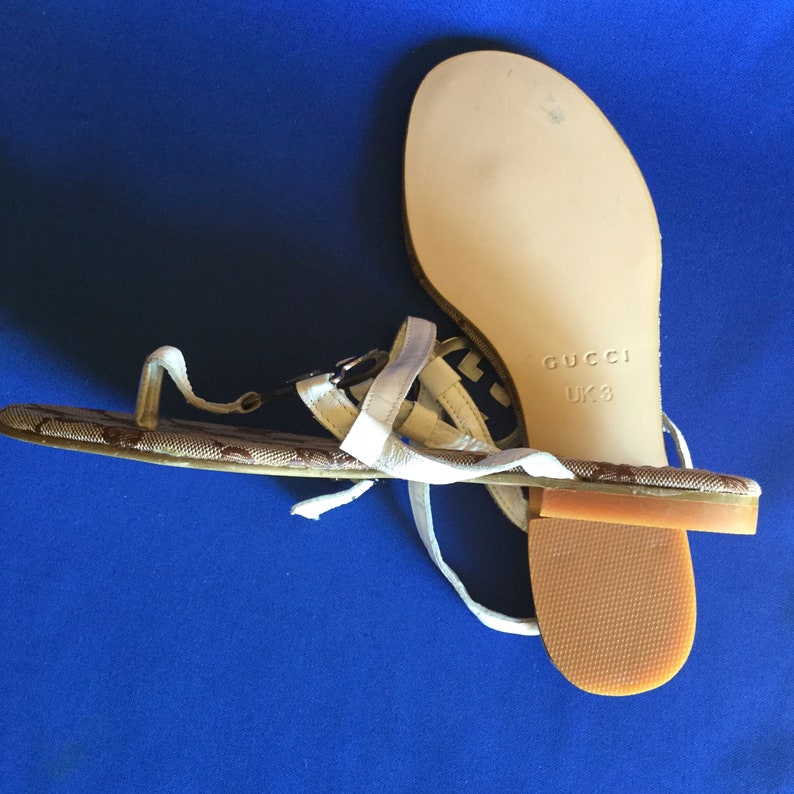 May include: White leather sandals with a brown and black patterned strap. The sandals have a brown sole and say "GUCCI UK 3" on the sole.