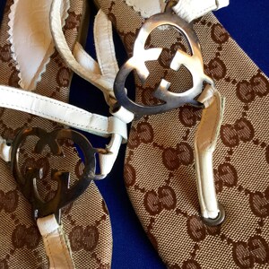 May include: A pair of brown and white sandals with a white leather strap and a silver heart-shaped buckle with the Gucci logo. The sandals have a brown and white patterned fabric.
