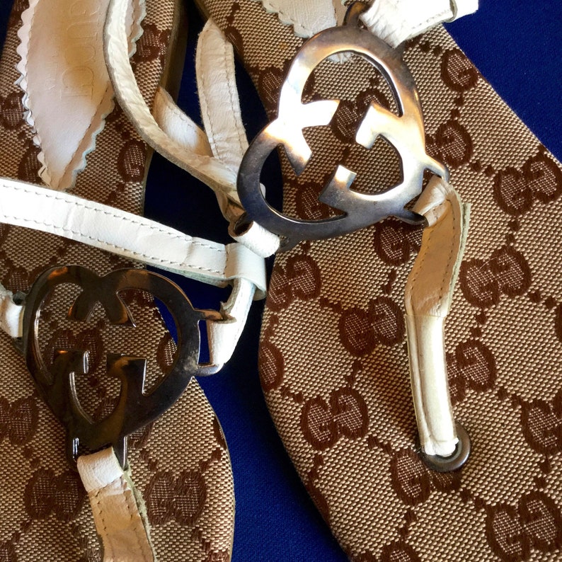 May include: A pair of brown and white sandals with a white leather strap and a silver heart-shaped buckle with the Gucci logo. The sandals have a brown and white patterned fabric.
