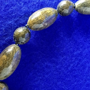 May include: A close-up of a necklace made of dark brown oval-shaped beads. The beads have a glossy finish and are strung together with black thread. The necklace is on a blue background.
