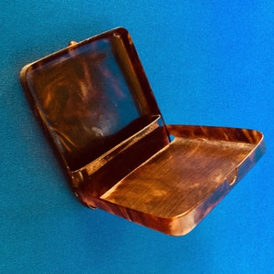 Puede incluir: Un estuche de cigarrillos de carey marrón con tapa con bisagras y un compartimento deslizable. El estuche está abierto y vacío.