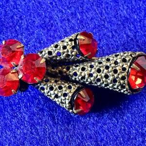 May include: A silver tone brooch with a cluster of red rhinestones. The brooch is shaped like a stylized flower with three long, pointed petals.