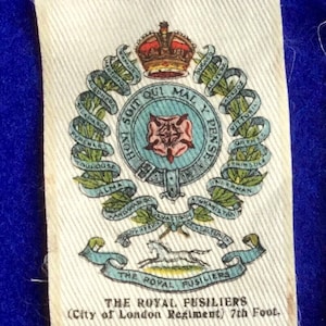Könnte beinhalten: Ein weißer Stoffabzeichen mit einem blauen und grünen Wappen, das eine Krone, eine Rose und ein Pferd zeigt. Der Text lautet "The Royal Fusiliers (City of London Regiment) 7th Foot. 108 B.D.V. Cigarettes."