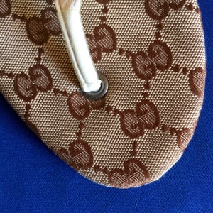 May include: Close-up of a brown and beige flip-flop with a white strap and a brown Gucci logo pattern.