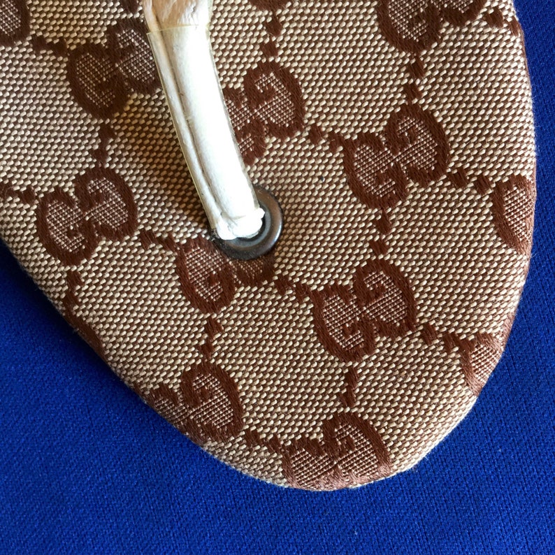 May include: Close-up of a brown and beige flip-flop with a white strap and a brown Gucci logo pattern.