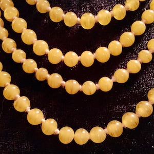 Puede incluir: Cuatro hilos de collares de cuentas amarillas sobre un fondo oscuro. Las cuentas redondas son translúcidas, con variaciones de color y textura. Los collares están ensartados en un cordón de color claro. Las cuentas miden unos 0,8 cm de diámetro.