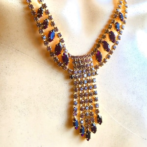 Könnte beinhalten: Eine goldene Halskette mit einem Anhänger aus violetten Strasssteinen. Die Kette hat eine lange, herabhängende Kette mit einem Cluster aus violetten Strasssteinen am unteren Ende.