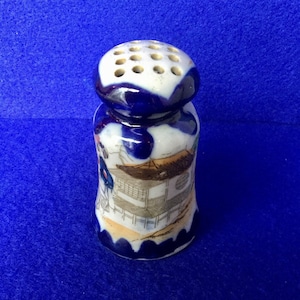 May include: A blue and white ceramic salt shaker with a Japanese design. The shaker has a round top with holes for sprinkling salt and a bell-shaped body with a scene of a building and figures.