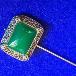 May include: A silver pin with a green stone set in a decorative frame. The pin is on a blue background.