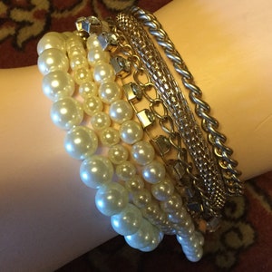 May include: A set of gold chain bracelets with a pearl strand bracelet. The bracelets are stacked on a person's wrist.