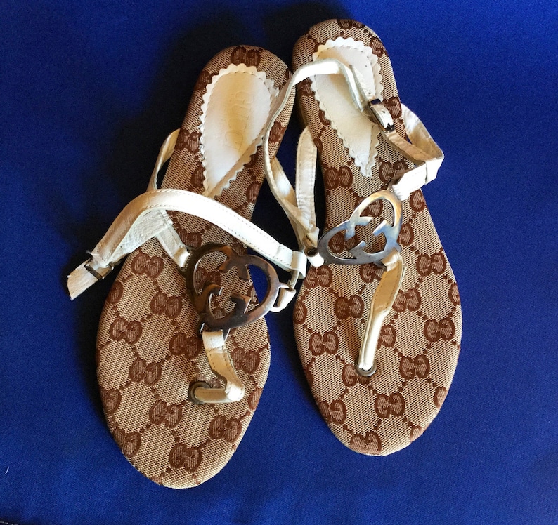 May include: A pair of white leather sandals with brown fabric straps and a gold GG logo. The sandals have a simple design and are perfect for summer.