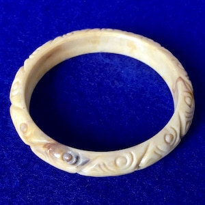 Può includere: Un braccialetto in avorio intagliato con un motivo decorativo a spirali e punti. Il braccialetto è di colore crema chiaro con una tonalità marrone leggermente più scura nei dettagli intagliati.