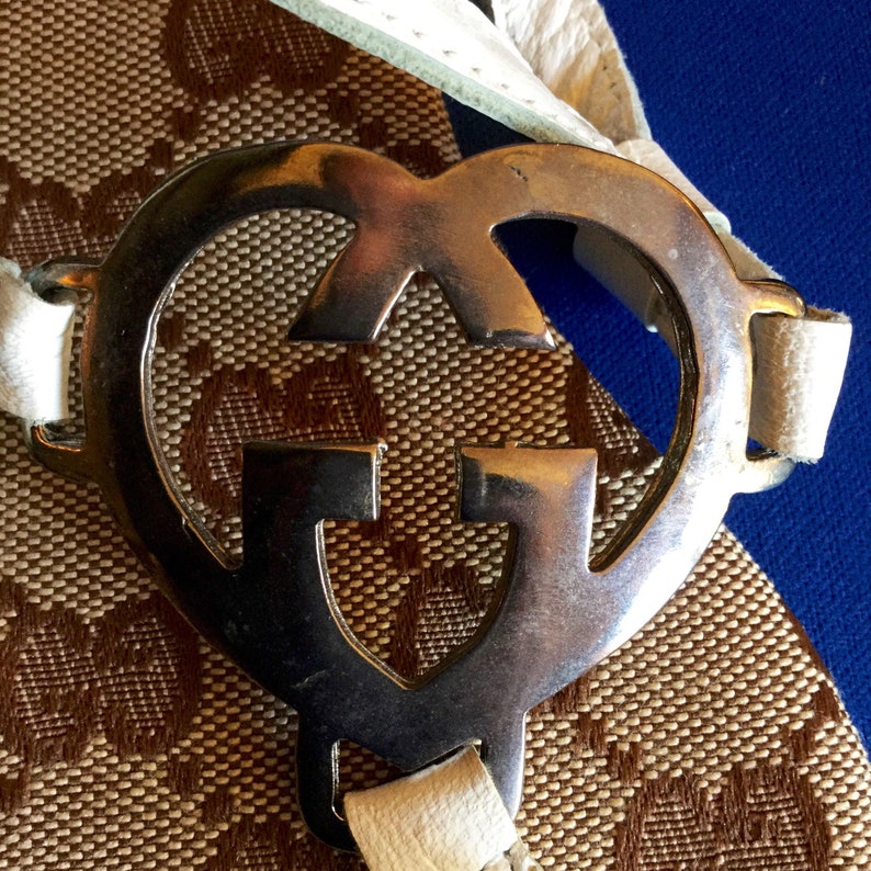 May include: A silver heart-shaped buckle with the Gucci logo in the center. The buckle is attached to a white leather strap.