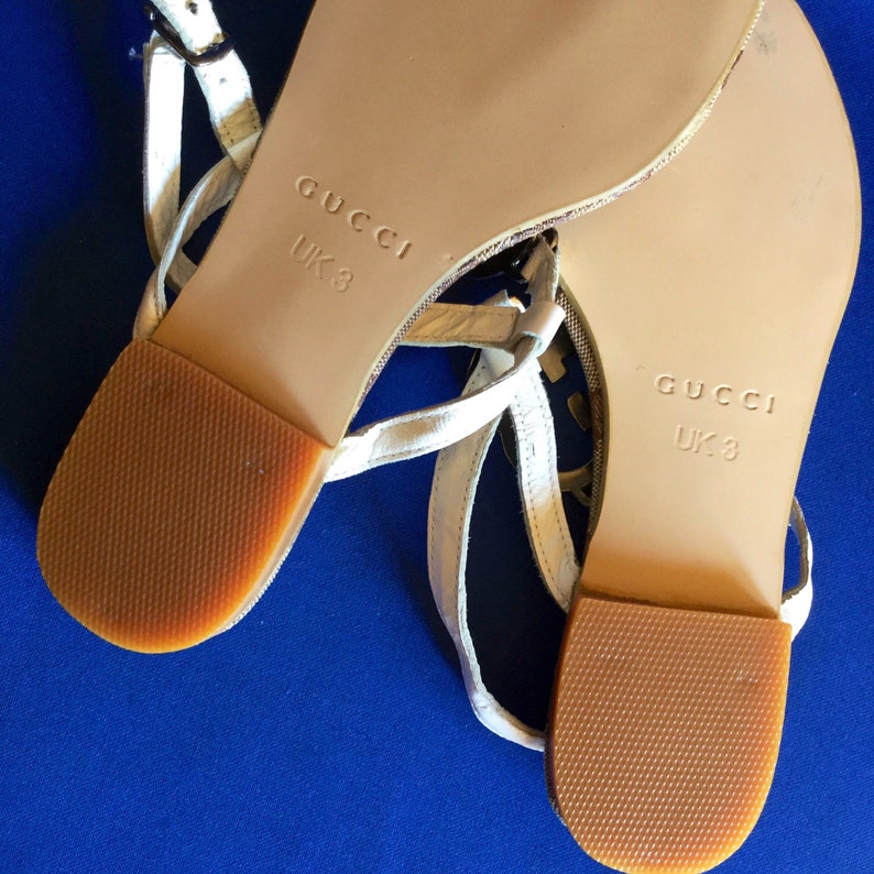 May include: White leather sandals with gold accents and brown soles. The sandals have the word "GUCCI" and "UK 3" printed on the soles.