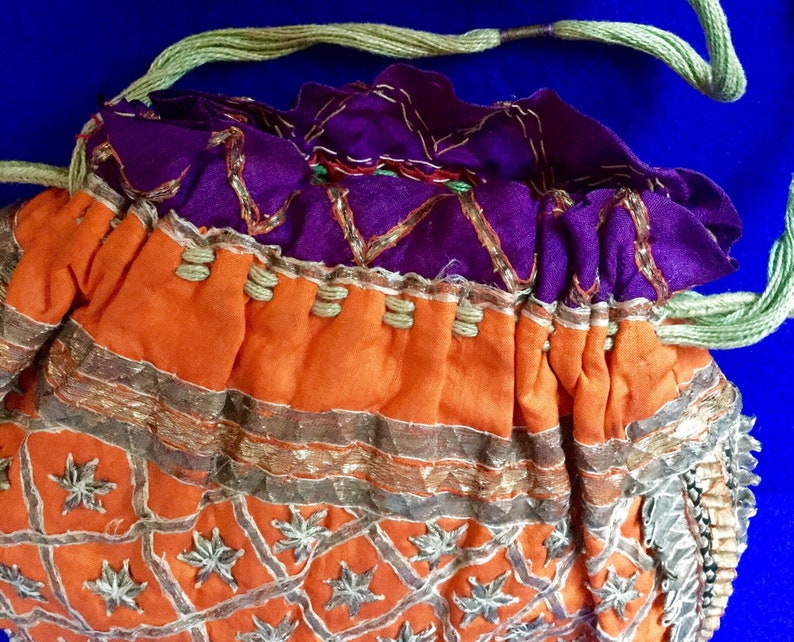 May include: A small, orange drawstring bag with gold and silver embroidery. The bag has a purple lining and a green drawstring.