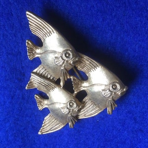 May include: Silver tone brooch featuring three stylized angelfish, each with detailed fins and eyes. The fish are arranged in a triangular shape.