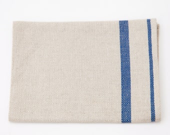 May include: Beige linen kitchen towel with two vertical blue stripes on the right side. The stripes have a diamond pattern. The towel is folded and lies flat on a white surface.