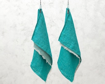 May include: Two turquoise linen hand towels with a fringed edge hang from metal hooks against a textured white background. The towels are folded in a triangle shape, showcasing the texture and color of the fabric.